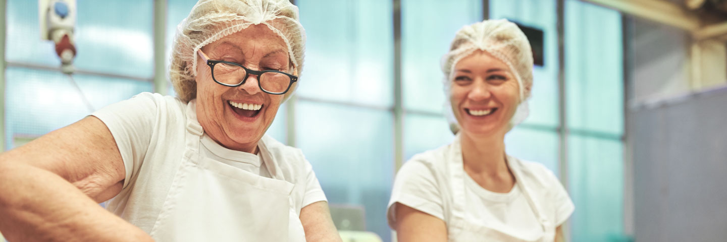 Eine ältere und eine jüngere Kollegin stehen in einer Fabrik lachend zusammen.