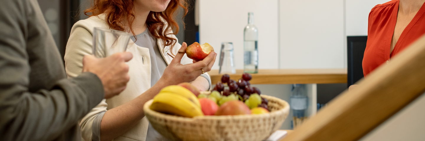 Kollegen stehen ein Obst essend um einen Obstkorb herum