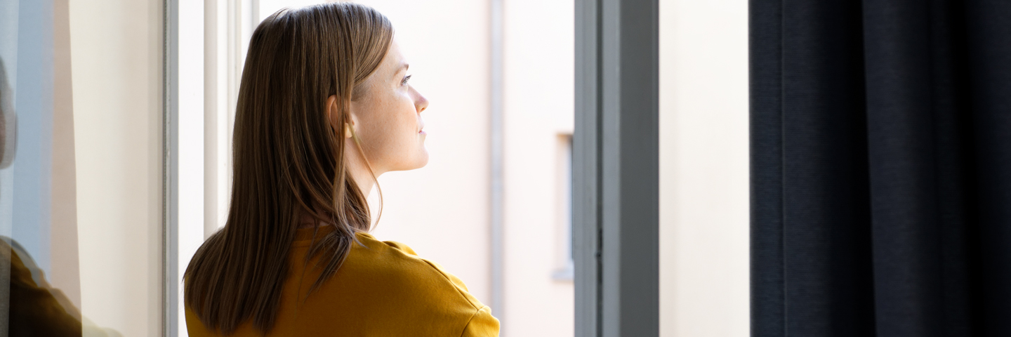 Junge Frau steht mit Blick in die Ferne am geöffneten Fenster.