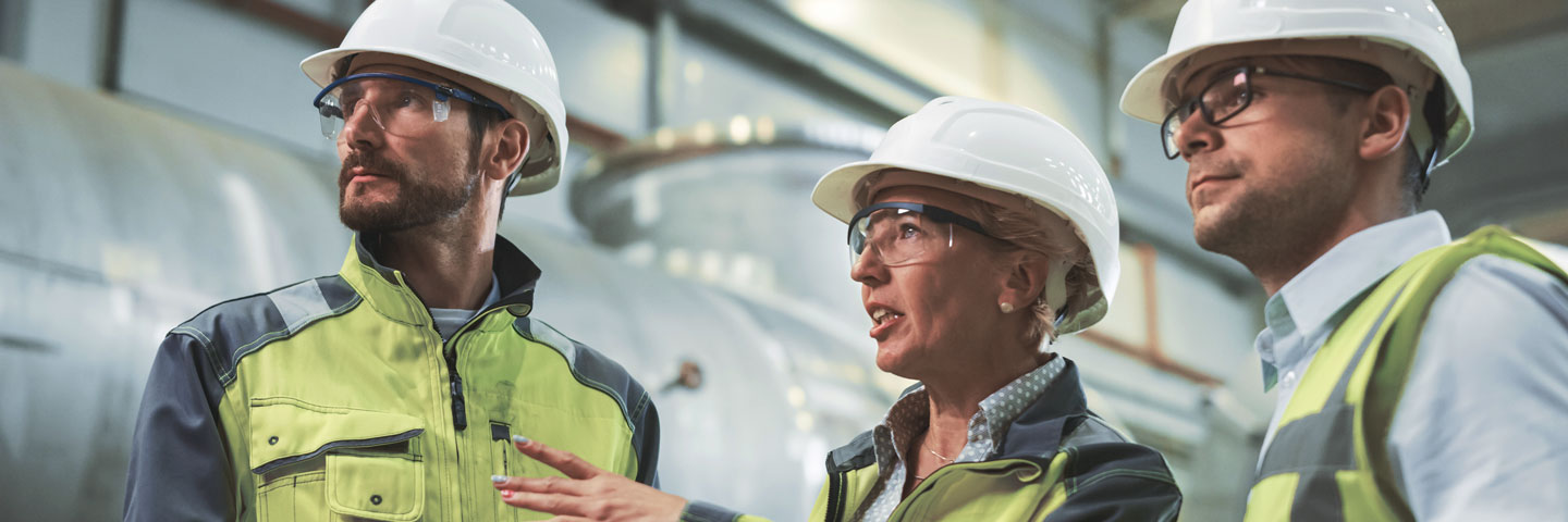 Zwei Männer und eine Frau in Sicherheitswesen, mit Schutzbrillen und Helmen stehen in einer Fabrikhalle.