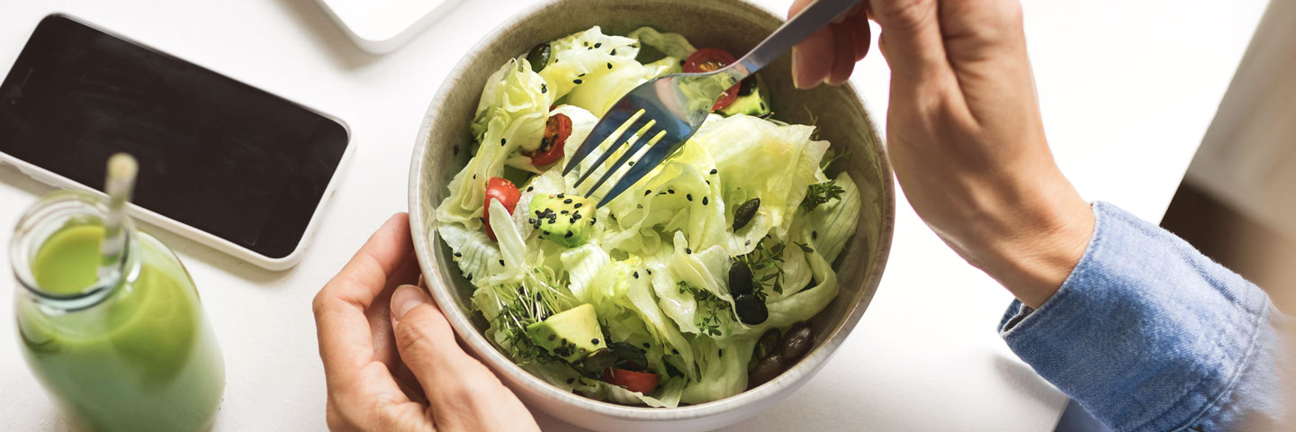 Zwei Hände, die einen Salat mit Dressing am Schreibtisch anrichten.