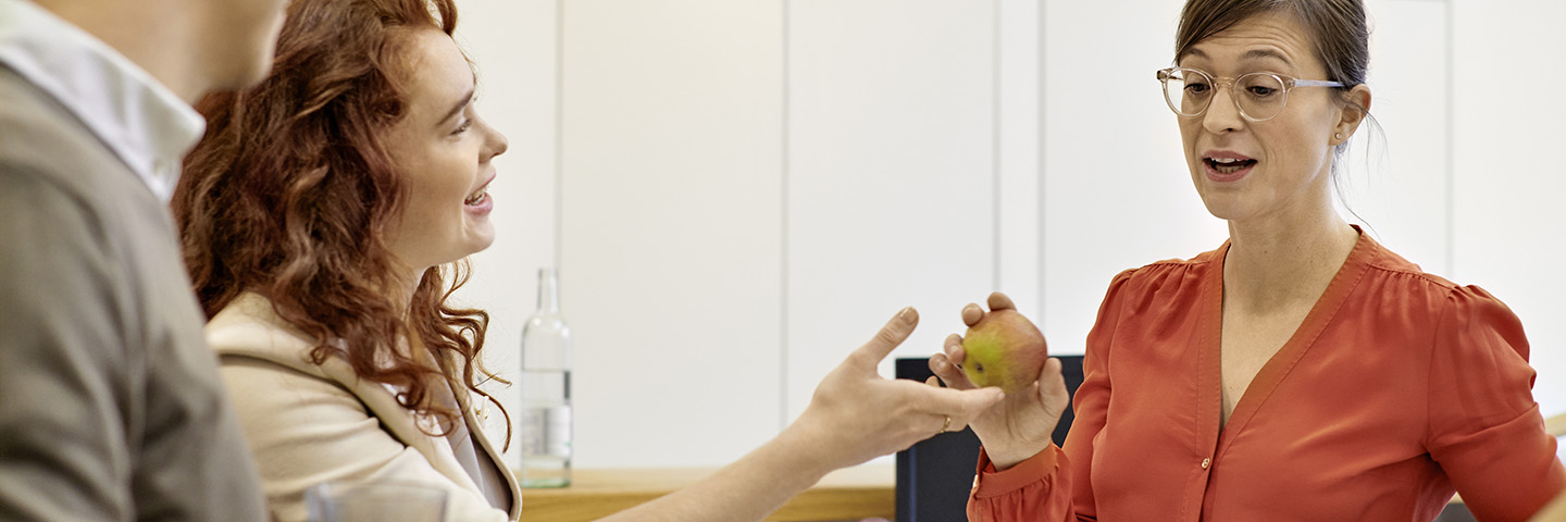 Arbeitgeber können mit Nudges gesundes Verhalten der Beschäftigten anstoßen. Drei Kollegen stehen zusammen um einen Obstkorb herum.