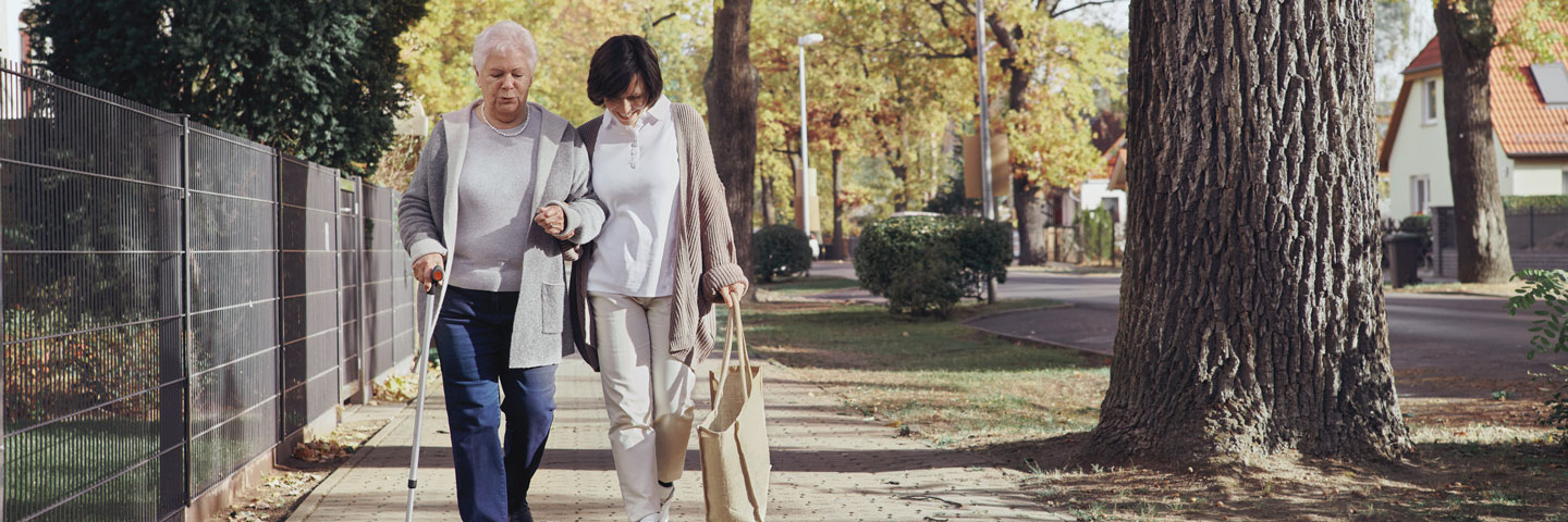 Eine Pflegekraft unterstützt eine Seniorin beim Spaziergang.