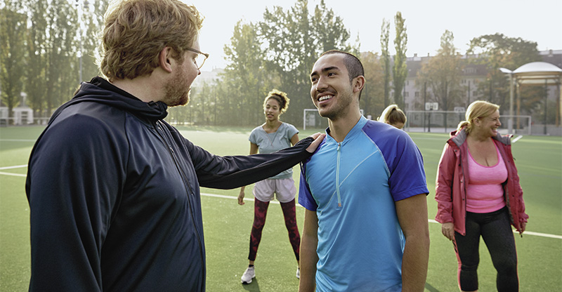 Zwei Männer stehen in sportlicher Kleidung auf einem Fußballfeld. Ein Mann legt seine Hand auf die Schulter des anderen Mannes. Im Hintergrund steht eine weitere PersonAOK.