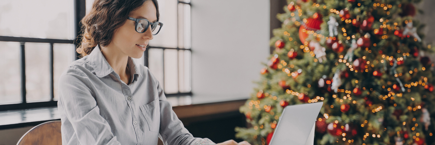 Digitaler Adventskalender Bayern: Eine lächelnde Frau mit Brille arbeitet an ihrem Laptop, während im Hintergrund ein festlich geschmückter Weihnachtsbaum mit roten Kugeln und Lichtern steht.