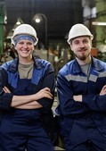 Frauen- und Männergesundheit im Betrieb Eine Frau und ein Mann in Arbeitskleidung und mit Schutzhelm stehen lächelnd mit verschränkten Armen in einer Werkhalle.