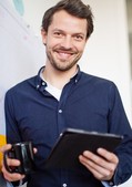 Online-Seminare für Arbeitgeber Lächelnder Mann mit Tablet und Kaffeetasse steht vor einem Whiteboard mit bunten Haftnotizen.