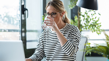 Eine Frau sitzt am Tisch und trinkt Wasser aus einem Glas.