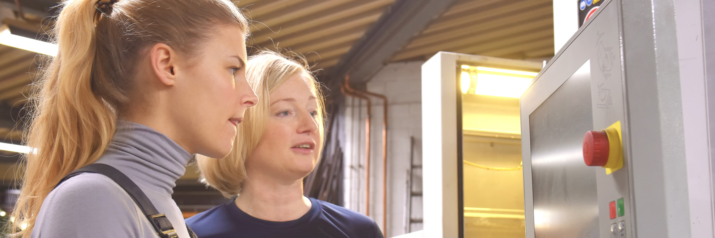 Onboarding für Fachkräfte: Eine junge Frau wird in einer Werkhalle angeleitet eine Steuerungsanlage zu bedienen Onboarding für Fachkräfte: Eine junge Frau wird in einer Werkhalle angeleitet eine Steuerungsanlage zu bedienen