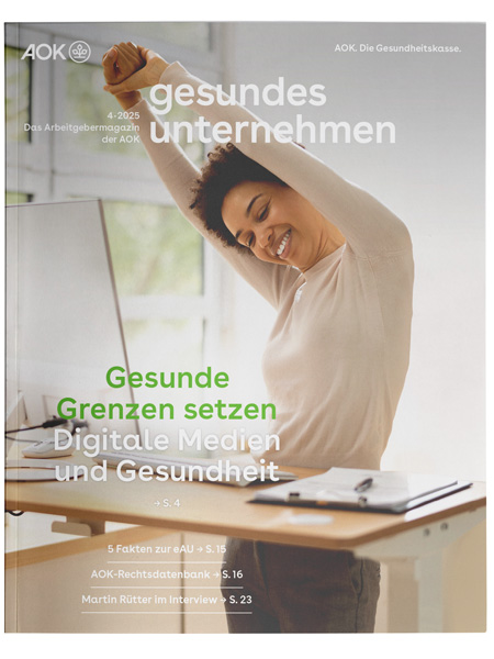 gesundes unternehmen Ausgabe 4-2025: Eine am Schreibtisch stehende Frau streckt sich gut gelaunt