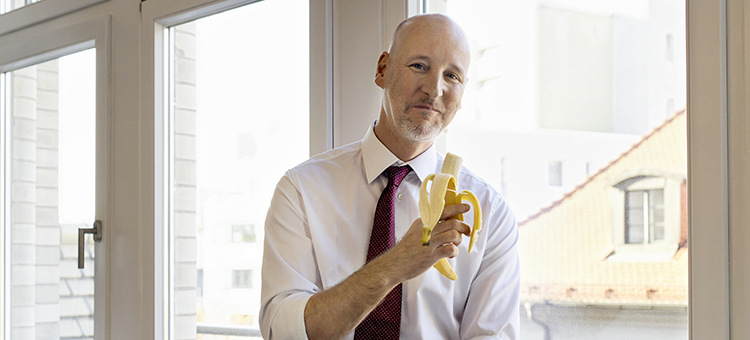 Schluss mit Stress-Essen im Job Ein Mann sitzt auf einem Bürofensterbrett und isst eine Banane.