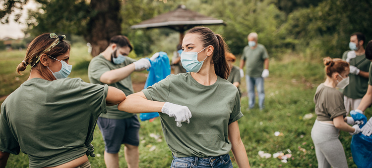 Gemeinsame Aktionen für den Umweltschutz stärken den Teamgeist. Zwei Frauen in der Natur tragen Mund-Nasenschutz und begrüßen sich coronakonform, indem sie ihre Ellenbogen zusammenführen.
