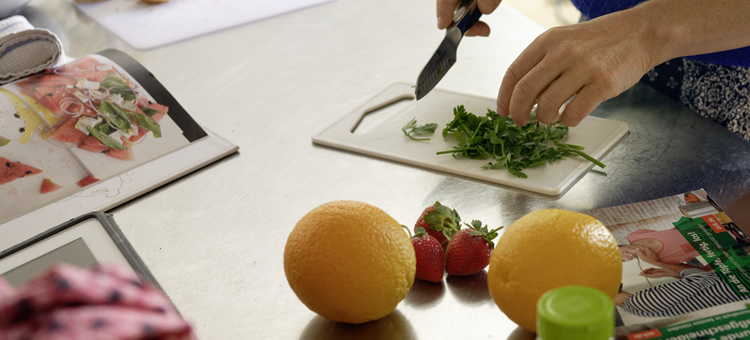 Wer vorkocht, kann sich im Arbeitsalltag gesünder ernähren. Jemand schneidet frische Kräuter, Obst liegt daneben auf dem Tisch.