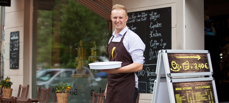 Minijobs: alte Zeitgrenzen und neue Geringfügigkeits-Richtlinien Junger Mann mit Schürze und Tablett vor einem Café.