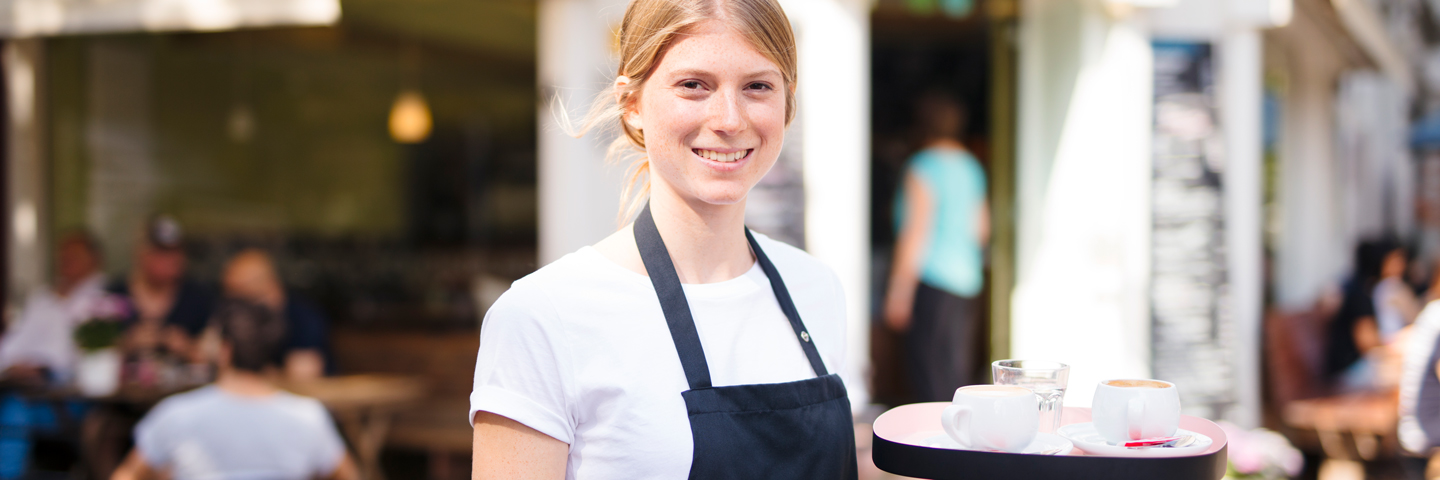 Eine junge Kellnerin steht bei Sonnenwetter im Außenbereich eines Gastronomiebetriebs.