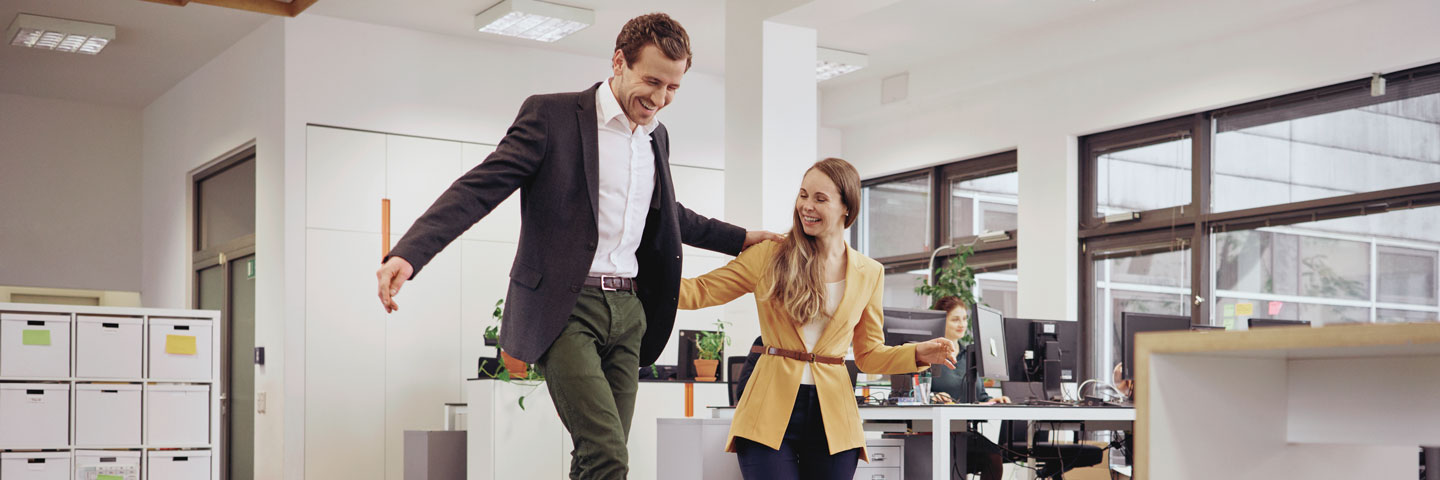 Ein Mann balanciert über eine Slackline im Büro. Eine Frau unterstützt ihn dabei. 