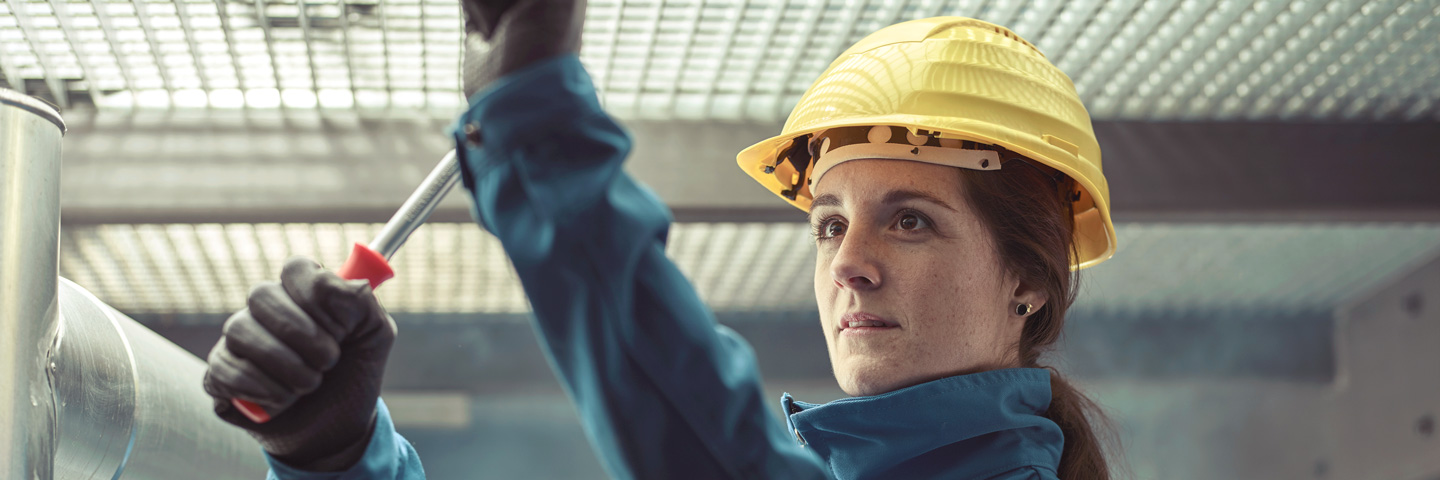 Kurzarbeit und Entgeltfortzahlung: Frau mit Schutzhelm und Schutzkleidung montiert eine Metallkonstruktion mit einem Schraubenzieher in einer Produktionshalle. Kurzarbeit und Entgeltfortzahlung: Frau mit Schutzhelm und Schutzkleidung montiert eine Metallkonstruktion mit einem Schraubenzieher in einer Produktionshalle.