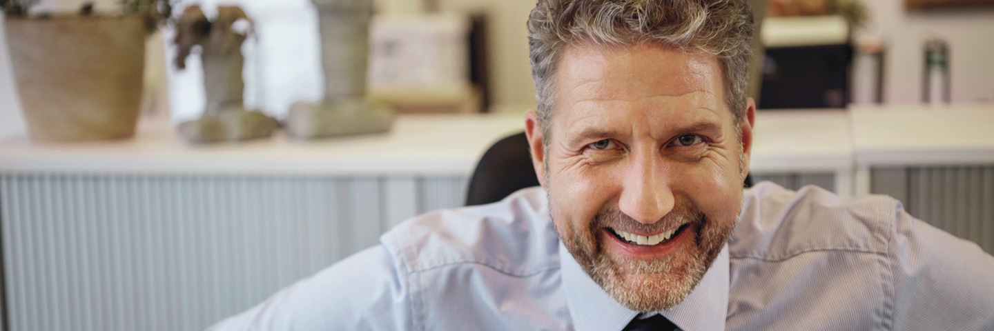 Resilienz im Arbeitsalltag: Lächelnder Mann im Büro sitzt am Schreibtisch und schaut in die Kamera