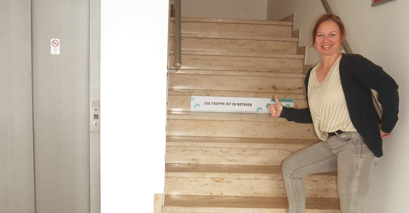 Frau steht im Treppenhaus und zeigt auf die Treppe mit dem Hinweis: Die Treppe ist in Betrieb. 