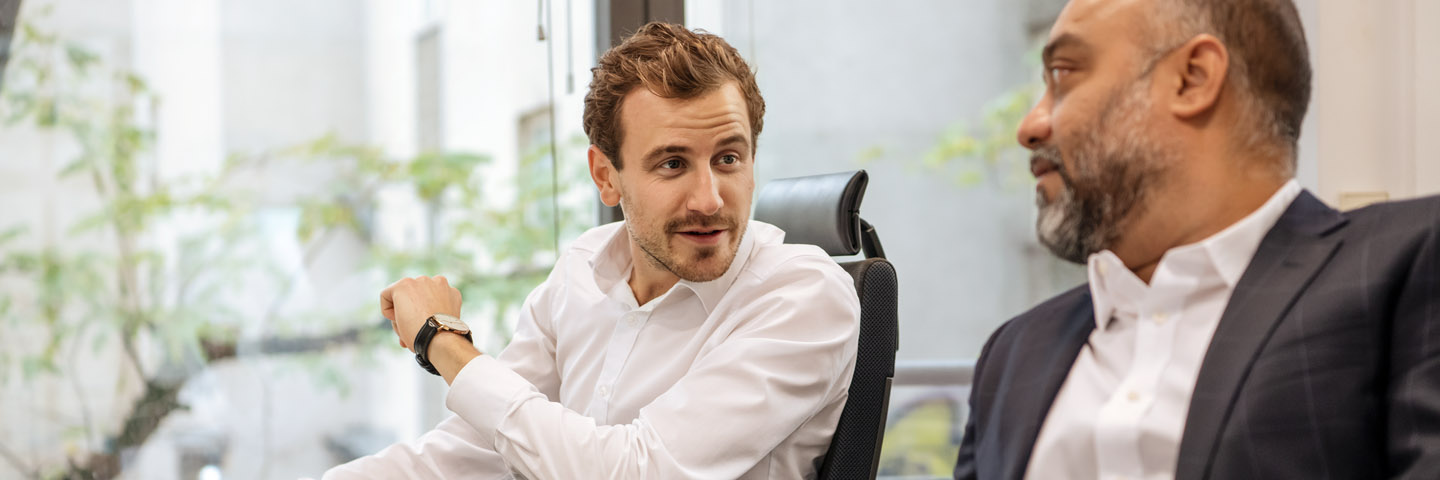 Zwei Männer sitzen nebeneinander im Büro und führen ein Gespräch. 