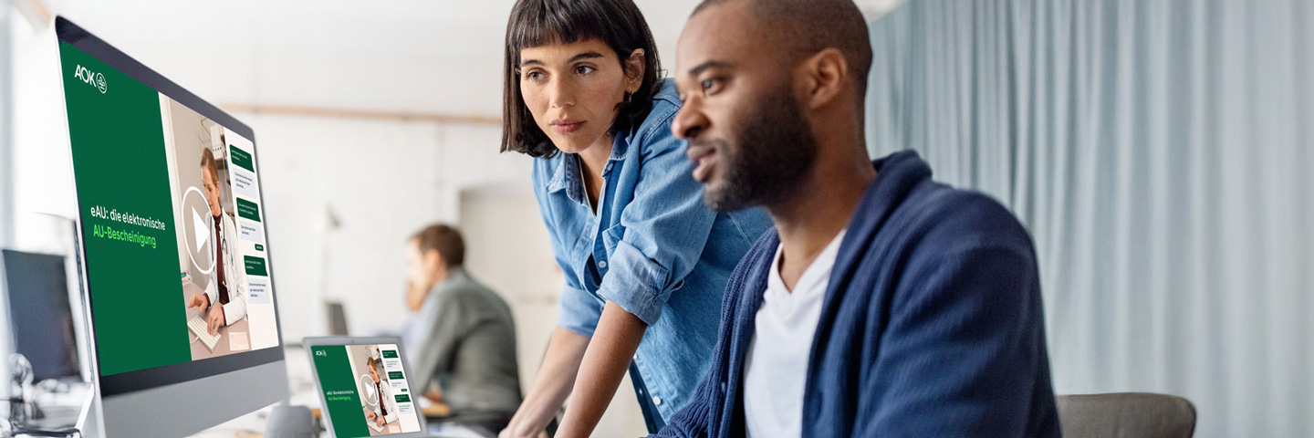 Seminar-on-demand für Arbeitgeber der AOK Zwei Personen schauen gemeinsam auf einen Monitor, auf dem ein Seminar-on-demand gezeigt wird.