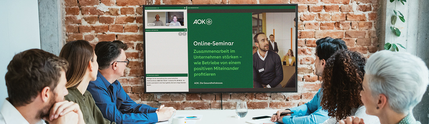 Eine Gruppe sitzt an einem Besprechungstisch und schaut sich auf einem Bildschirm an der Rückwand des Raumes die Aufzeichnung eines Online-Seminars an.