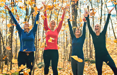 Gesund durch den Herbst: Eine Gruppe sportlich gekleideter Frauen wirft in einem Wald fröhlich Laub hoch.