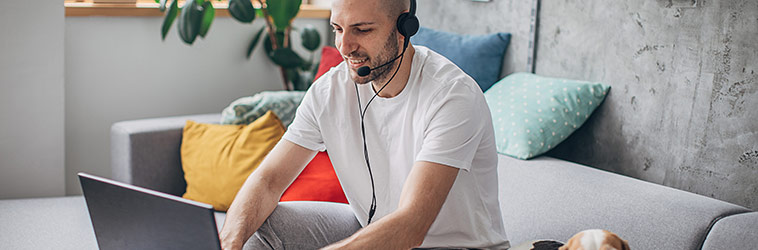 Online-Seminar „#marketplace – gesund gründen“ Existenzgründer sitzt auf dem heimischen Sofa mit Notebook und Headset freudig bei der Arbeit, Hund schaut in die Kamera.