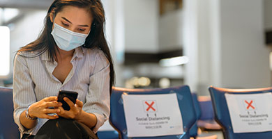 Eine Frau mit Mund-Nasen-Schutz wartet im Flughafen auf ihren Flug und schaut auf ihr Smartphone.