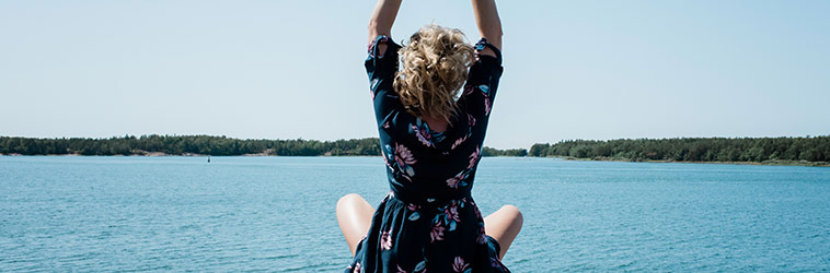 Online-Seminar „Resilienz“ Frau sitzt an einem See und streckt sich tief durchatmend.