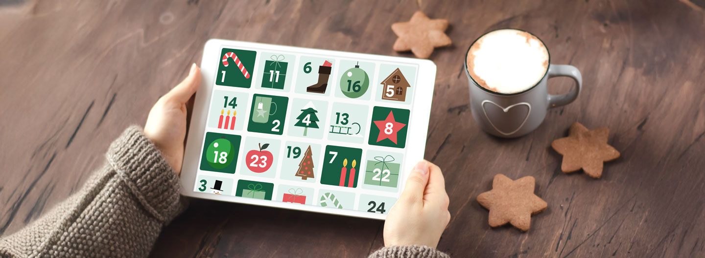 Eine Person hält ein Tablet, das einen digitalen Adventskalender anzeigt. Daneben sind eine Tasse Kaffee und Plätzchen auf einem Holztisch zu sehen.