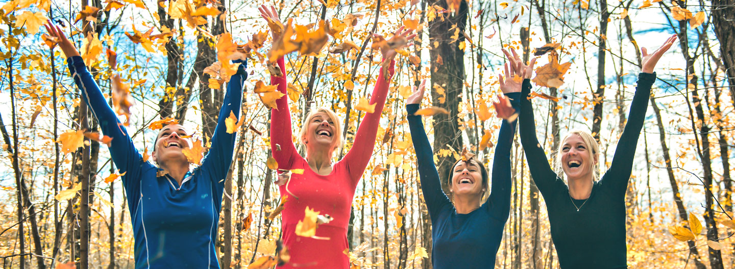 Gesund durch den Herbst: Eine Gruppe sportlich gekleideter Frauen wirft in einem Wald fröhlich Laub hoch.