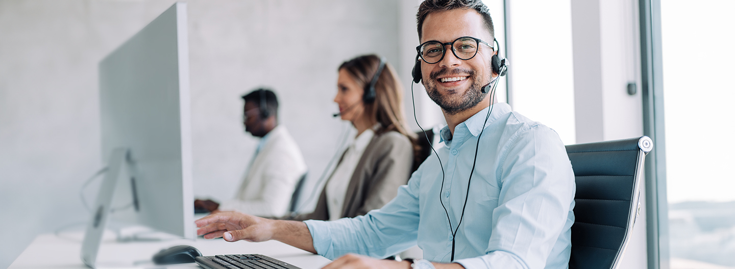 Drei Personen mit Headset sitzen an einem Schreibtisch. Ein Mann lächelt dabei freundlich in die Kamera.