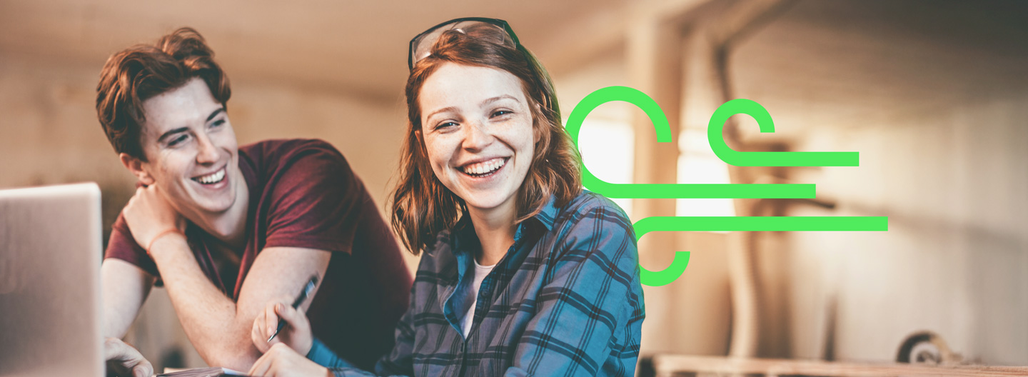 Zwei lächelnde Auszubildende in einer Werkstatt-Umgebung mit eingeblendetem grünen Grafik-Symbol.