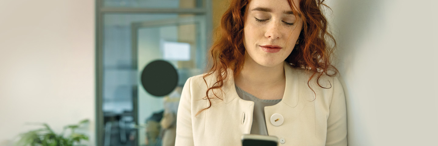Beiträge zur Sozialversicherung Rothaarige Frau im Büro schaut auf ihr Smartphone.