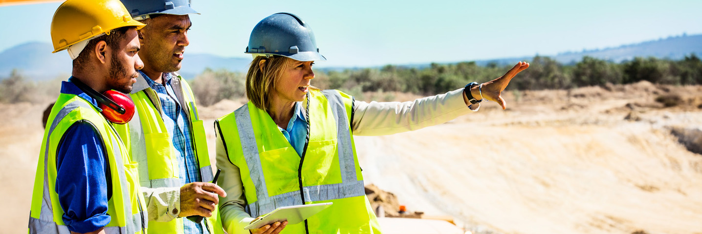 Entsendungen ins Ausland in der Sozialversicherung Eine Frau und zwei Männer mit Helm und Weste besprechen sich auf einer Baustelle.