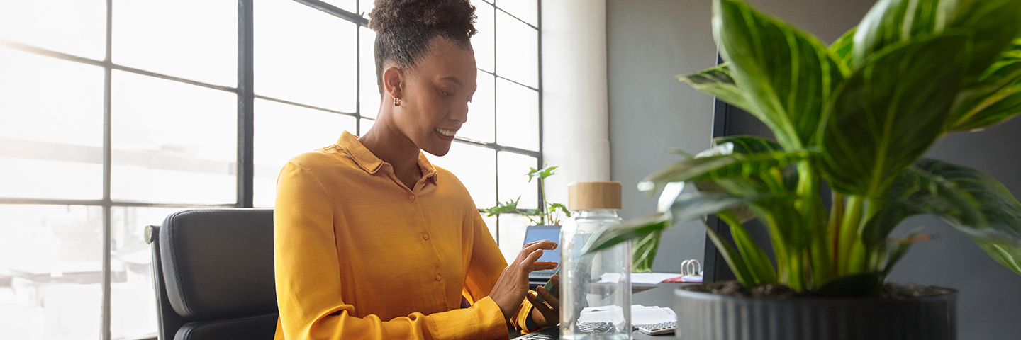 Eine Frau sitzt an einem Schreibtisch in einem hellen Büro und blickt konzentriert auf ein Smartphone in ihrer Hand, vor ihr eine Tastatur und ein Computermonitor. 