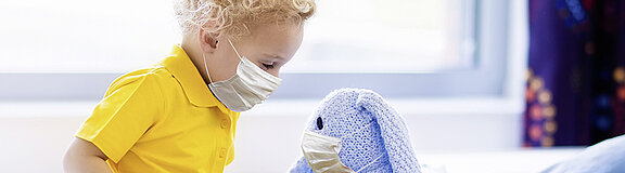 Ein kleiner Junge, der einen Mundschutz trägt, sitzt im Krankenhausbett und schaut auf sein Kuscheltier.