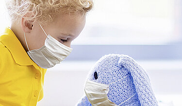Ein kleiner Junge, der einen Mundschutz trägt, sitzt im Krankenhausbett und schaut auf sein Kuscheltier.