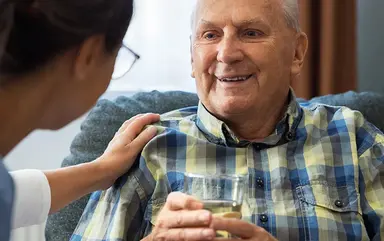 Pflegekraft unterstützt einen älteren Mann mit einem Glas Wasser in einer Pflegeeinrichtung.