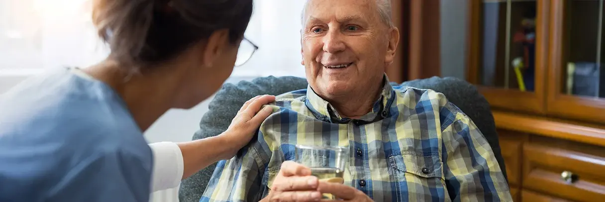 Pflegekraft unterstützt einen älteren Mann mit einem Glas Wasser in einer Pflegeeinrichtung.