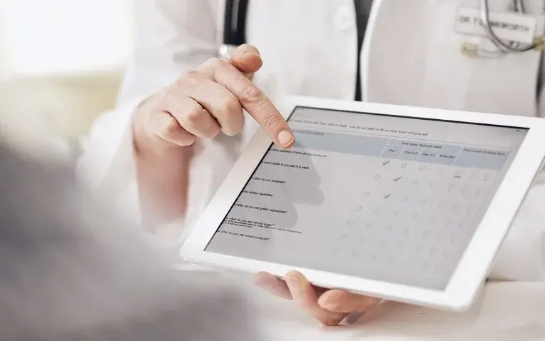 Arzt zeigt auf Tablet mit Fragebogen zur Krankenhauszufriedenheit, Patient im Vordergrund unscharf