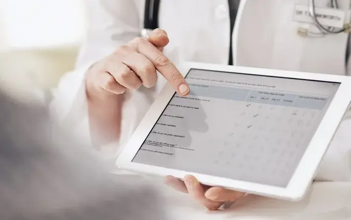 Arzt zeigt auf Tablet mit Fragebogen zur Krankenhauszufriedenheit, Patient im Vordergrund unscharf