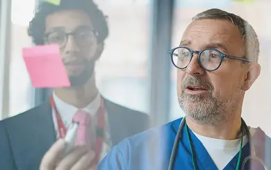 Mehrere Personen in medizinischer Kleidung stehen vor einer Glaswand mit bunten Haftnotizen und diskutieren.