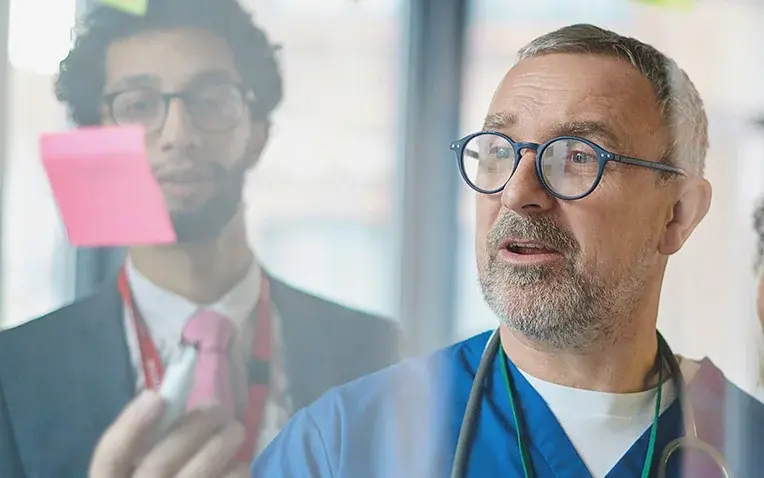 Mehrere Personen in medizinischer Kleidung stehen vor einer Glaswand mit bunten Haftnotizen und diskutieren.