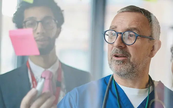 Mehrere Personen in medizinischer Kleidung stehen vor einer Glaswand mit bunten Haftnotizen und diskutieren.