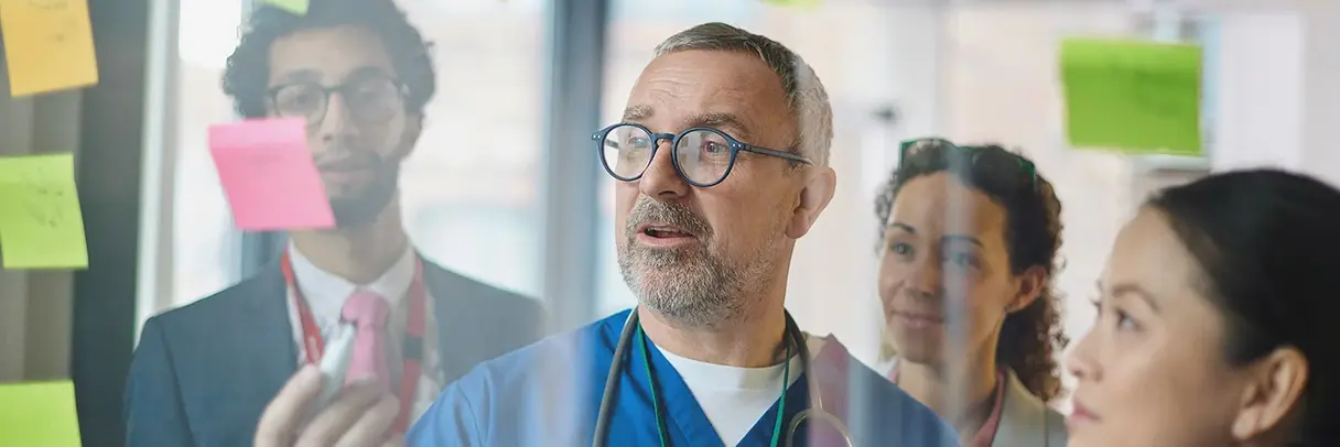 Mehrere Personen in medizinischer Kleidung stehen vor einer Glaswand mit bunten Haftnotizen und diskutieren.