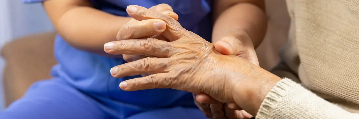 Eine Pflegekraft untersucht die Hand einer älteren Person – Fokus auf geschwollene Finger und Gelenke, möglicherweise durch rheumatoide Arthritis verursacht.