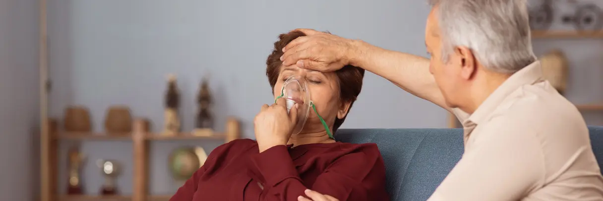 Ein älterer Mann untersucht den Kopf einer sitzenden Person in einem roten Hemd, vermutlich in einem Wohnzimmer mit Regalen im Hintergrund. Das Bild illustriert einen Beitrag zum QISA-Band F3 „Gesundheitsversorgung im Klimawandel“. 