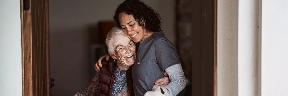 Eine Pflegekraft in Arbeitskleidung umarmt eine ältere Frau im Türrahmen. Beide lächeln.